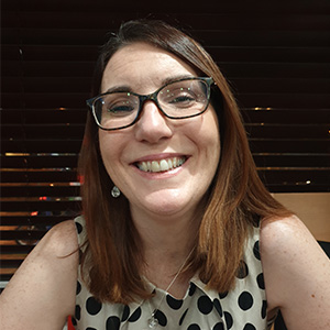 Photo of the head and shoulders of Elaine Arundell, a white female with long brown hair and dark rimmed glasses. Elaine is wearing a sleeveless white dress with black polka dots.