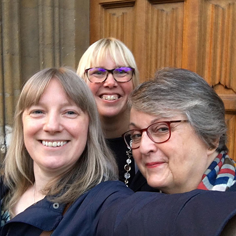 Three ladies standing in front of a wooden church door. All of them are smiling and looking at the camera. On the left is a lady with shoulder length light brown straight hair and a fringe, her left arm is outstretched in order to take the selfie. In the middle at the back is a blond haired lady wearing brown glasses. On the right is a grey-haired lady wearing red glasses. She has a red, white and blue scarf around her neck.