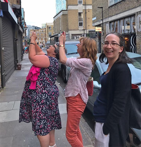 Three ladies stood on the pavement in an urban area. Two of them are facing a building and holding their mobile phones up to take a phot of the building. A third lady is in the foreground looking at the camera and smiling