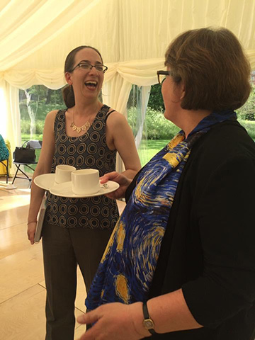 Two ladies in a marquee in a garden on a sunny day. The lady on the left has long dark hair in a ponytail and is wearing a blue sleeveless top with grey and dark blue circles and black trousers. She is holding two plates in her left hand with a cup balanced on each one. In het left hand she is holding a notebook. She is. in conversation with the lady on the right who is facing away from the camera. She has short brown hair and is wearing a patterned blue and yellow top and a black cardigan.