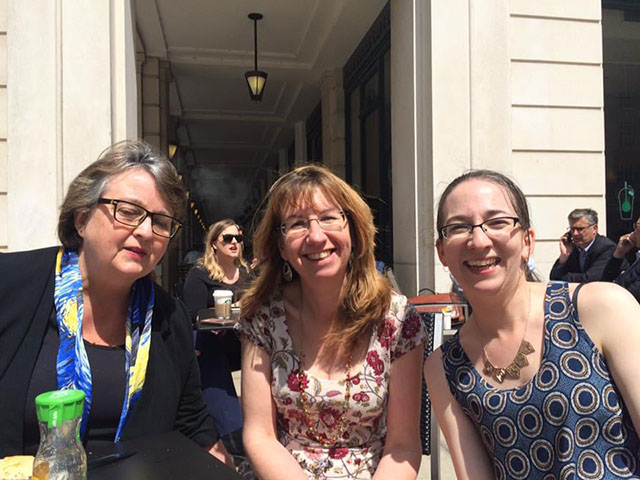 Three ladies sat on a terrace of a cafe. They are sat in a line facing the camera. The lady on the left has short brown hair and is wearing glasses. She is dressed in a dark blue top with a blue and yellow patterned scarf around her neck. The lady in the middle has long light brown hair and is wearing glasses. She has a flowery short sleeved dress on .The lady on the right has long dark hair in a ponytail and is wearing a blue sleeveless top with grey and dark blue circles 
