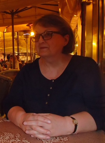 A lady with short brown hair and brown glasses sitting at a table in a dimly lit pub. She has her hands clasped together on the table in front of her and is looking sideways at the camera.