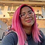 A black woman with long pink hair, wearing clear glasses and a grey short sleeved t-shirt. She is stood outside a building, smiling and looking at the camera.